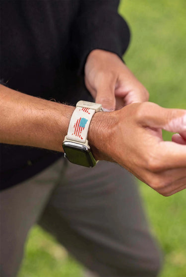 A close-up of a person's wrist wearing a smartwatch with a cream-colored band printed with a red, white, and blue American flag design, set against a blurred green outdoor background.