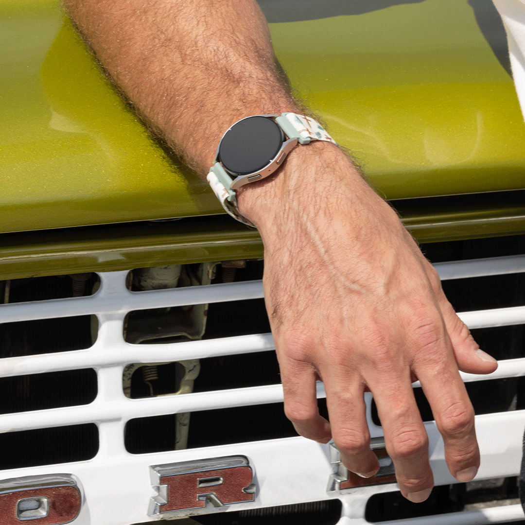 A close-up of a person's hand resting on the front grille of a vintage car while wearing a smartwatch with a light green and white patterned strap.