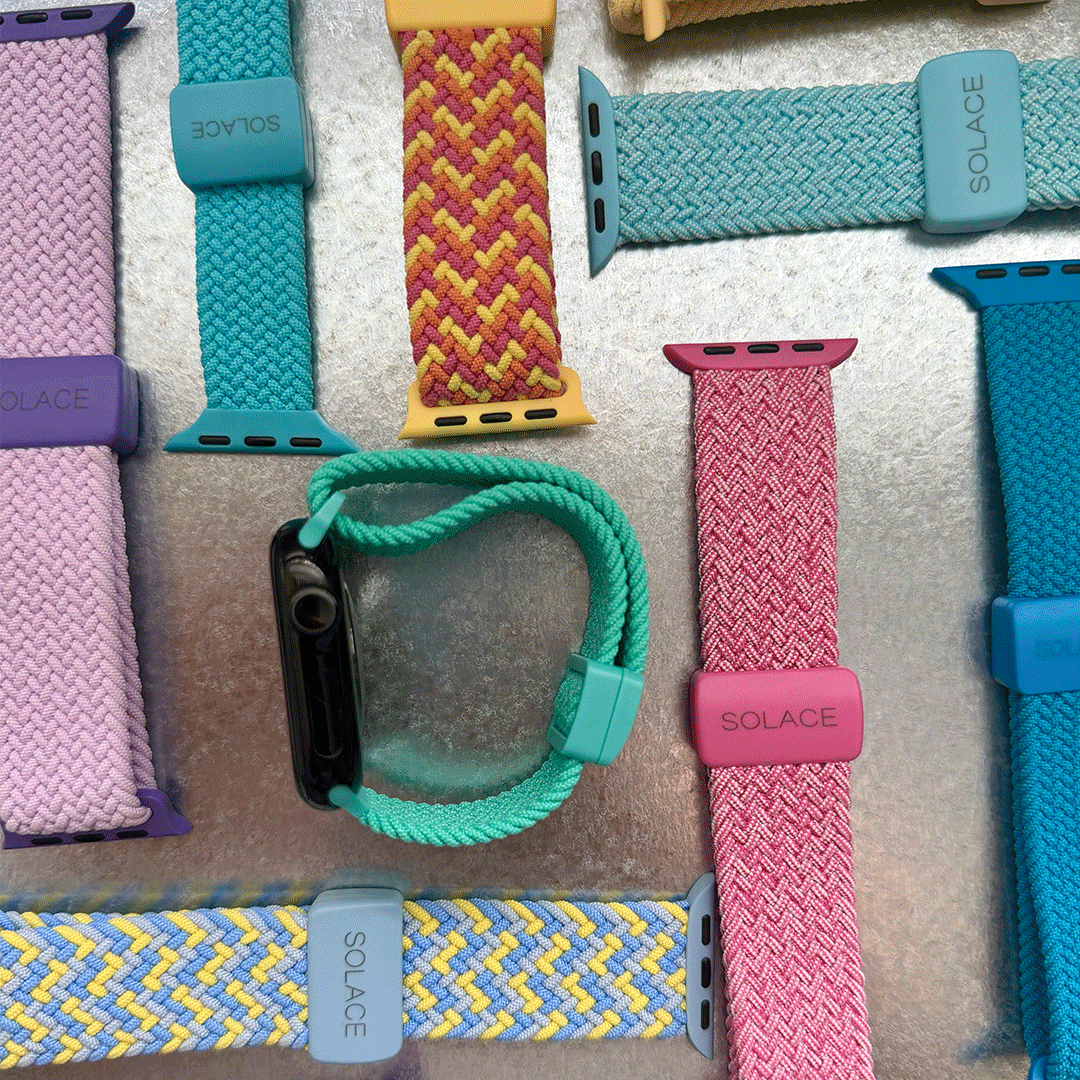 A collection of colorful braided silicone watch straps by SOLACE, shown laid out on a metallic silver surface. The straps feature various colors including teal, pink, yellow, blue, and purple, with one teal strap shown attached to a black smartwatch in the center.