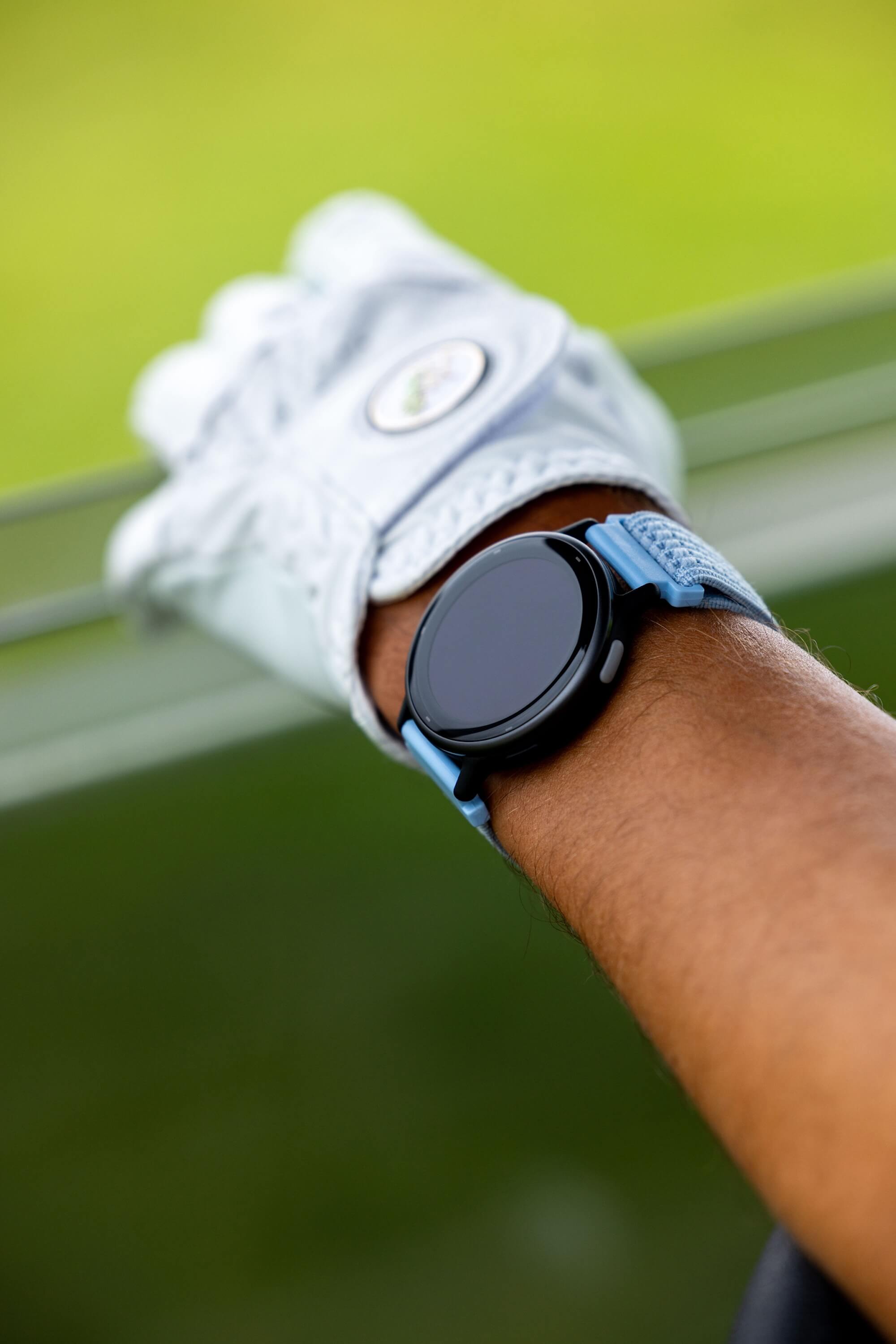 A close-up of a smartwatch with a light blue and black strap worn on a person's wrist, with a white golf glove visible in the background against a blurred green outdoor setting.