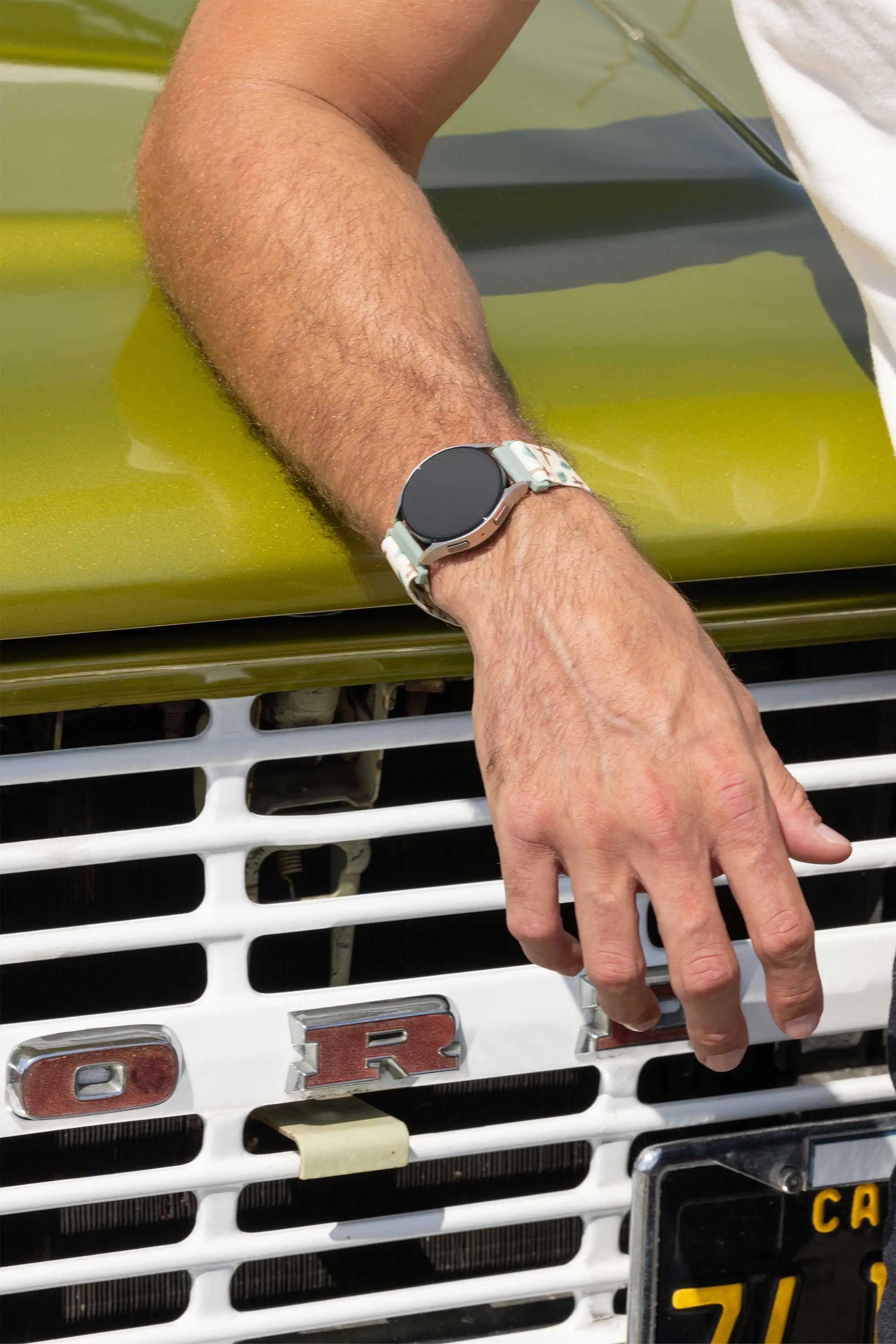 A close-up lifestyle shot of a person's hand resting on the chrome grille of a vintage lime green car, wearing a silver smartwatch with a white woven strap.