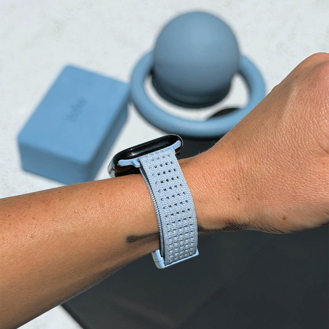 A close-up of a person's wrist wearing a light blue textured sport band attached to a smartwatch, with a matching light blue yoga/exercise ball and a small blue box in the background on a light surface.