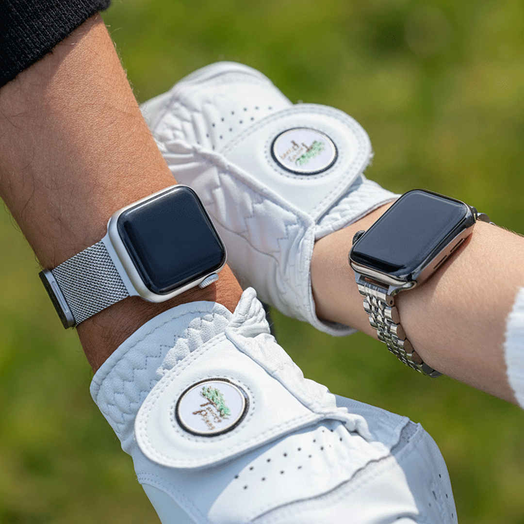 Two wrists wearing white golf gloves and smartwatches, photographed outdoors with a blurred green grass background. The golf gloves have circular white logos with green and gold detailing.